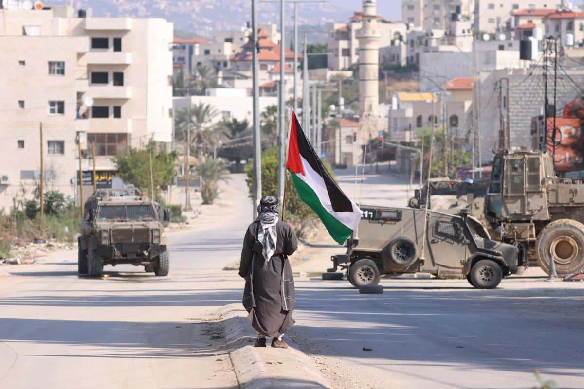 Un hombre lleva una bandera palestina mientras vehículos militares israelíes avanzan por una calle en la ciudad cisjordana de Tulkarem.